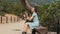 Young resting woman with dreadlocks sitting on the bench near the tree and using her phone