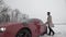 Young rich man walking on snowy field to red muscle car