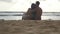 Young romantic couple is enjoying beautiful view sitting on the beach and hugging at evening. A woman and a man sits