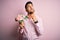 Young romatic man holding bouquet of spring flowers over pink isolatd background serious face thinking about question, very