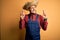 Young rural farmer man wearing bib overall and countryside hat over yellow background gesturing finger crossed smiling with hope