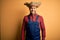 Young rural farmer man wearing bib overall and countryside hat over yellow background with a happy and cool smile on face