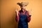 Young rural farmer man wearing bib overall and countryside hat over yellow background shouting with crazy expression doing rock