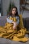 Young sad girl sitting on a comfortable couch, covered with a yellow blanket, clasped her knees with her hands