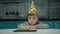 Young sad preschooler boy in birthday hat looking to candle on piece of cake. Lonely upset birthday kid sitting at home