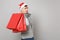 Young Santa man in Christmas hat holding red package bag with gifts, purchases after shopping on grey