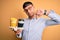 Young scholarship holder business man holding take away coffee over isolated yellow background with angry face, negative sign