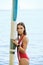 Young serene brunette woman in red swimwear standing by her surfboard