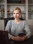 Young serious adorable blonde business woman posing in home office room sitting at the desk with notebook holding mobile phone