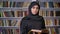 Young serious muslim girl in hijab is reading book, watching at camera, religious concept, bookshelf on background