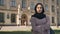 Young serious muslim girl is standing with arms crossed in daytime in summer, watching at camera, building on background