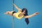 Young sexy brunette making superman element hanging on pole at blue background. Fit smiling poledancer in yellow pole