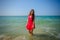 Young sexy long-haired brunette in red beach dress stands in the turquoise water of the ocean on a hot day. Beautiful smiling