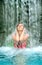 Young sexy woman in pink bikini holds hands on head, enjoy the falling water of the waterfall in the spa pool, copy space