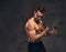 Young shirtless bodybuilder with muscular body doing exercise with dumbbells on dark background.