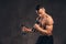 Young shirtless bodybuilder with muscular body doing exercise with dumbbells on dark background.