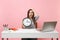 Young shocked woman clinging to head hold alarm clock while sit work at office with pc laptop isolated on pastel pink