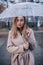 A young, slender, beautiful girl in a coat walks in the rain with a transparent umbrella in the park