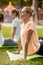 Young slender girl with closing eyes doing yoga exercises on the yoga mat with other girls on green grass in the park on