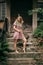 Young slender girl in short dress made of natural fabric is standing on steps of authentic house with fern in hands