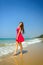 Young slender long-haired brunette in red dress standing barefoot on tropical beach with yellow sand against the sea and blue sky