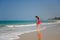 Young slender long-haired brunette in red dress walks barefoot on tropical beach along the surf against the sea and blue sky