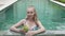 The young slender woman in bikini drinks coconut milk from a coco on the edge of the pool in the tropical resort