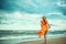 A young slender woman in orange dress is walking barefoot towards the storming sea