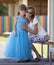 Young slim blond smiling mother, aunt or sister hugs small pretty preschool daughter girl in long nice blue evening dress on blur