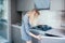 Young slim blonde woman in grey dress standing by the stove in the modern grey kitchen