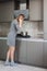 Young slim blonde woman in grey dress standing by the stove in the modern grey kitchen