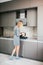 Young slim blonde woman in grey dress standing by the stove in the modern grey kitchen