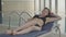 Young slim Caucasian woman in black bikini resting on sunbed in front of swimming pool in spa hotel. Portrait of slender