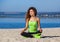 Young slim girl with curly hair in black and light green tracksuit sitting after a workout in the sand on the seashore.