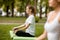 Young slim girl sits in the lotus position with closing eyes doing exercises with other girls on green grass in the park