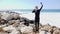 Young slim sportive happy man standing on the rocky beach taking selfies and showing thumbs up while water splashing agains the cl