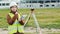 Young smileing woman surveyor in green work clothes and helmet adjusts the equipment, produces calculations and writes