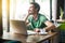 Young smiley happy businessman in green t-shirt sitting, working on laptop and looking outside, dreaming or pondering