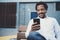 Young smiling african man using smartphone to listen to music while sitting on the bench at sunny street.Concept of