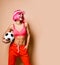 Young smiling athletic woman with pink hair in bright sportswear standing and holding soccer ball in hand