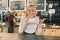 Young smiling barista girl with cup of coffee. Blonde woman in apron enjoying the aroma of fresh coffee, near a bar counter in