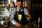 Young smiling barman decorates cocktail with ice in glass by mint petals.