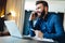 Young smiling bearded businessman sitting in front of computer, talking on cell phone, holding pen. Phone conversations.