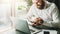 Young smiling bearded businessman in white shirt sitting at desk in front of laptop, making notes in notebook.