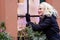 A young, smiling, blond woman from outside looks through a window adorned with Christmas decorations. The concept of Christmas