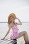 Young smiling blonde girl look like a Barbie doll in pink mini dress and wide brim hat sitting on beach