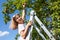 Young smiling girl stretches hand to hanging red apple, standing on ladder leaned against tree stem