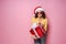 Young smiling happy cheerful curly girl in Santa& x27;s hat holds red giftbox while stands on pink background. Sopping