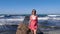 Young smiling happy woman walking on a sea pier barefoot. Red dress flapping on the wind. Waves hitting the sea pier.