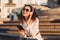 Young smiling lady with dark curly hair in sunglasses and white jacket sitting on stairs with cellphone in hand and